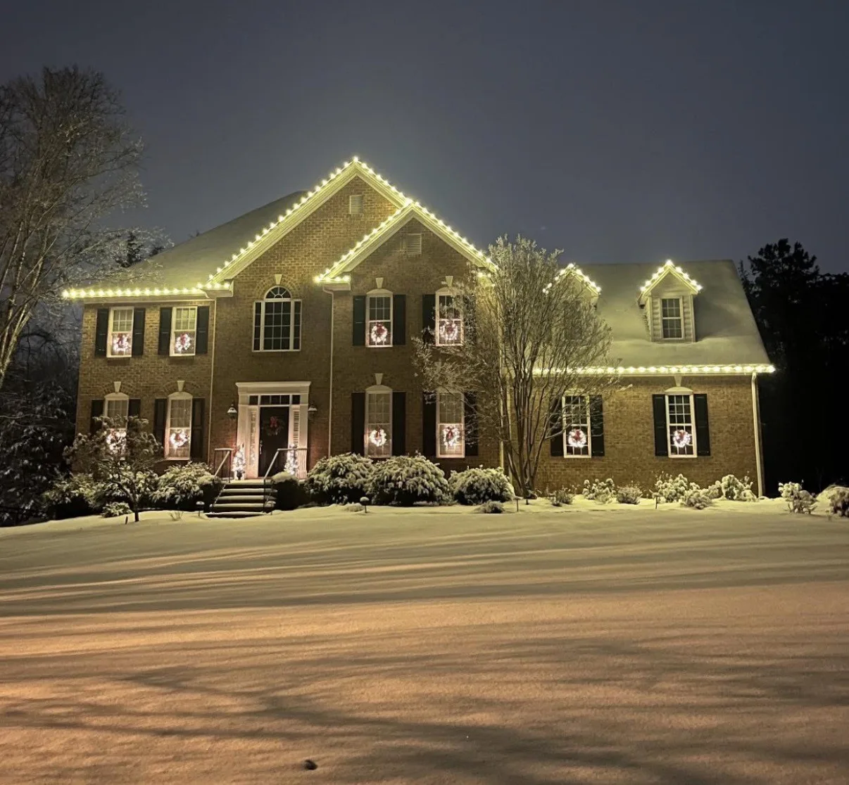 Two-story brick home on a snowy night with Christmas lights along the roofline, peaks and dormers and lit wreaths in the windows