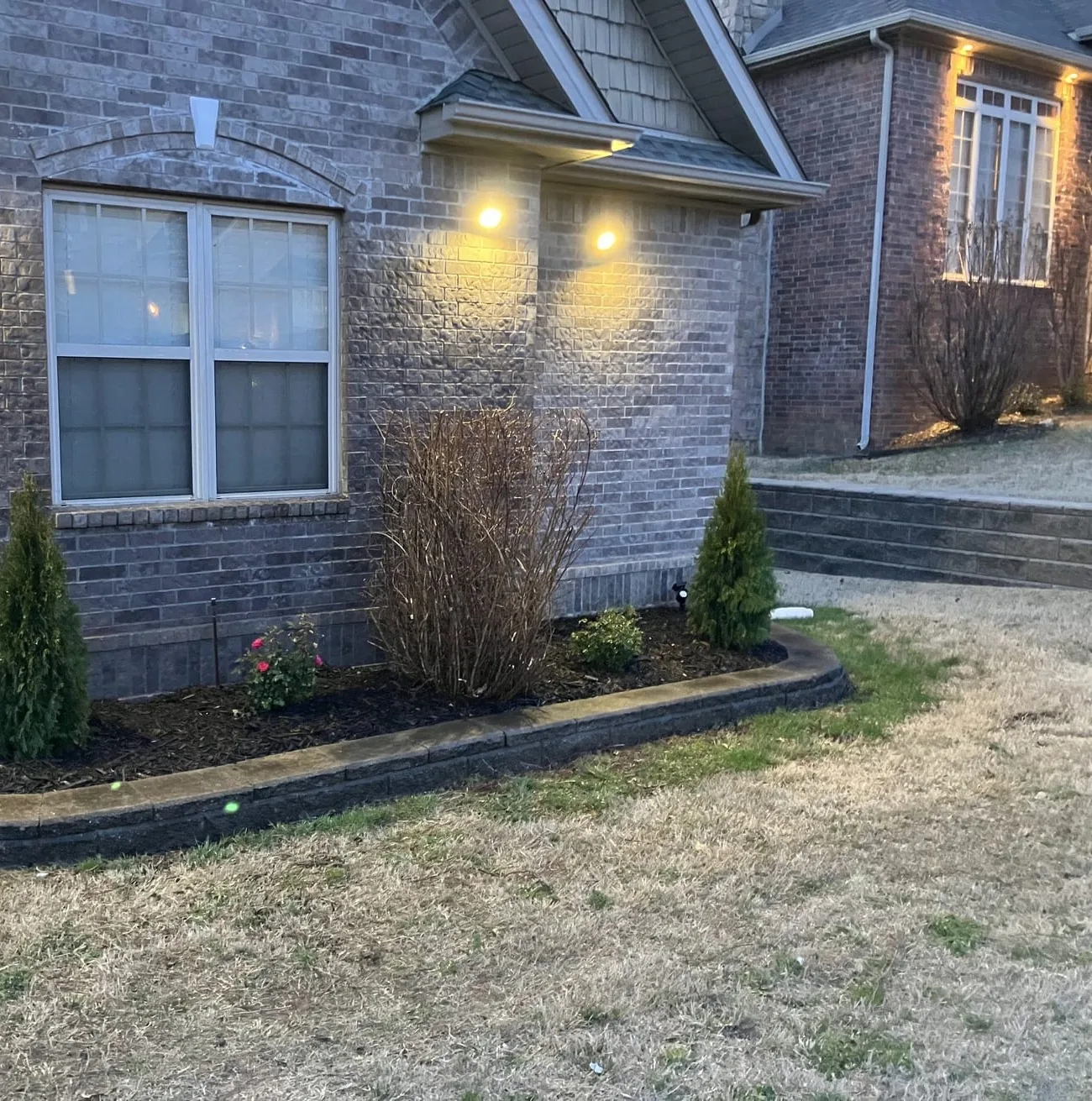 Brick home at dusk with lights washing the wall above a curbed front bed of young evergreens and shrubs