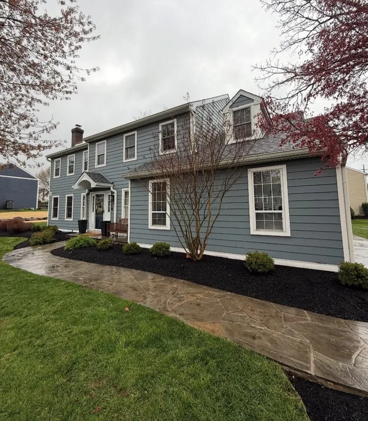New mulched planting bed with young shrubs along a paver walkway in front of a two-story home